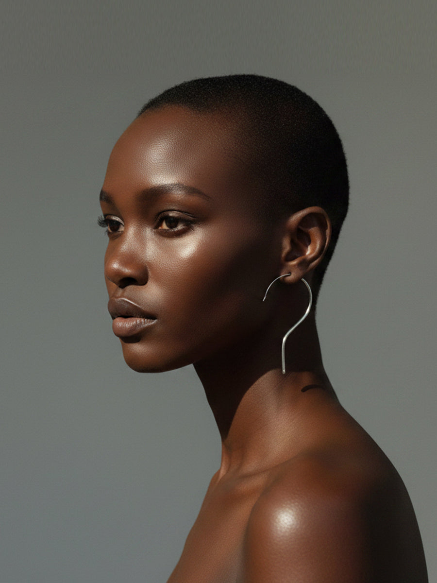 A close-up jewellery portrait of a woman in profile wearing a large, minimalist silver hanger earring against a solid gray studio background.