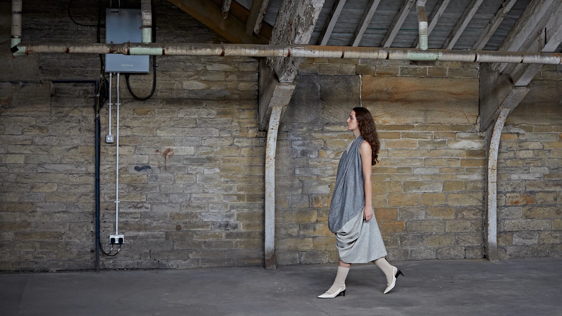 A model walks through a historic stone Yorkshire mill interior wearing a draped grey Cunnington & Sanderson garment, evoking the British craftsmanship and heritage behind every piece.