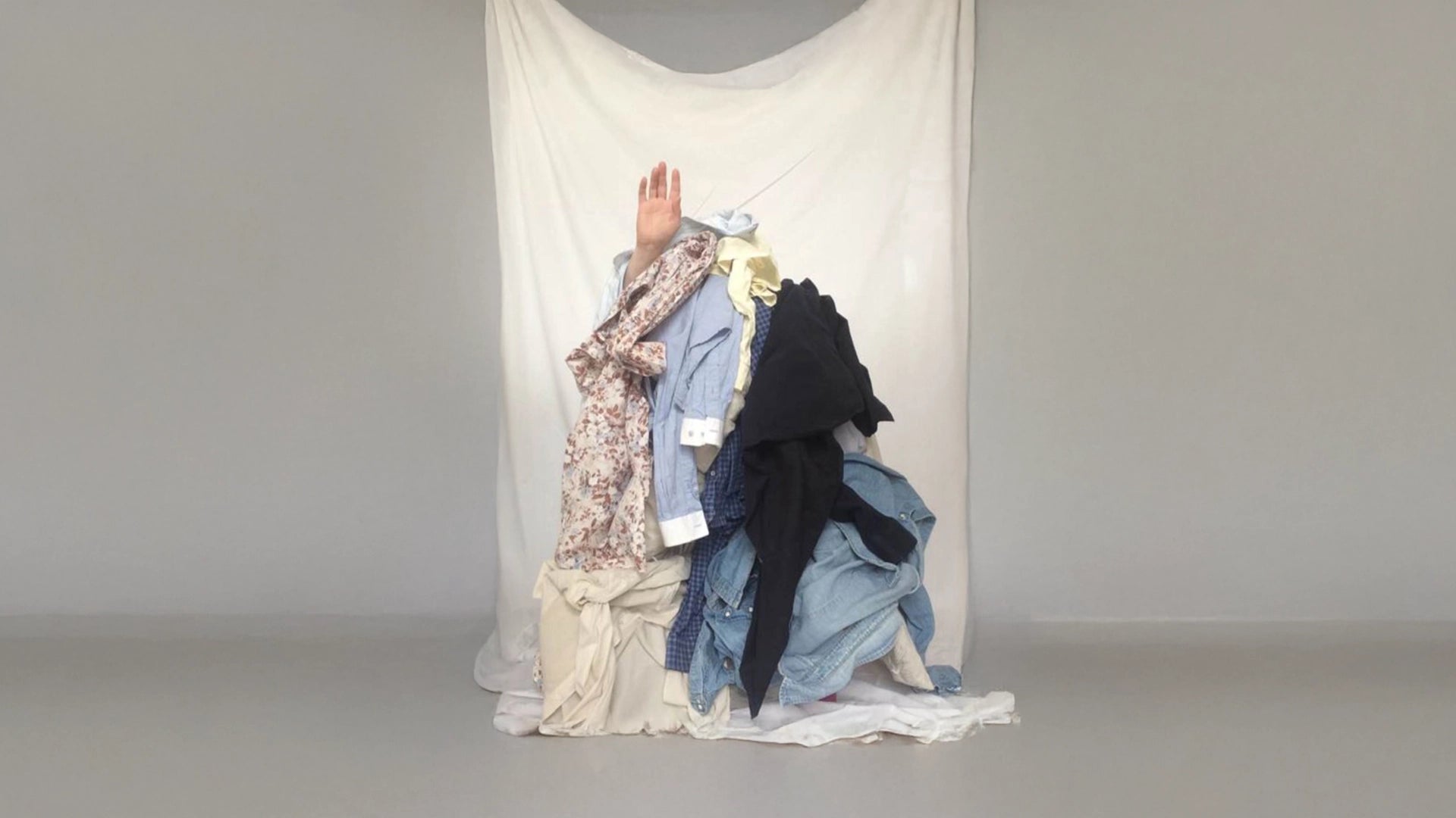 A pile of mixed clothing in floral, denim and navy against a draped cream backdrop, with a hand emerging from within — representing Cunnington & Sanderson's considered approach to materials.