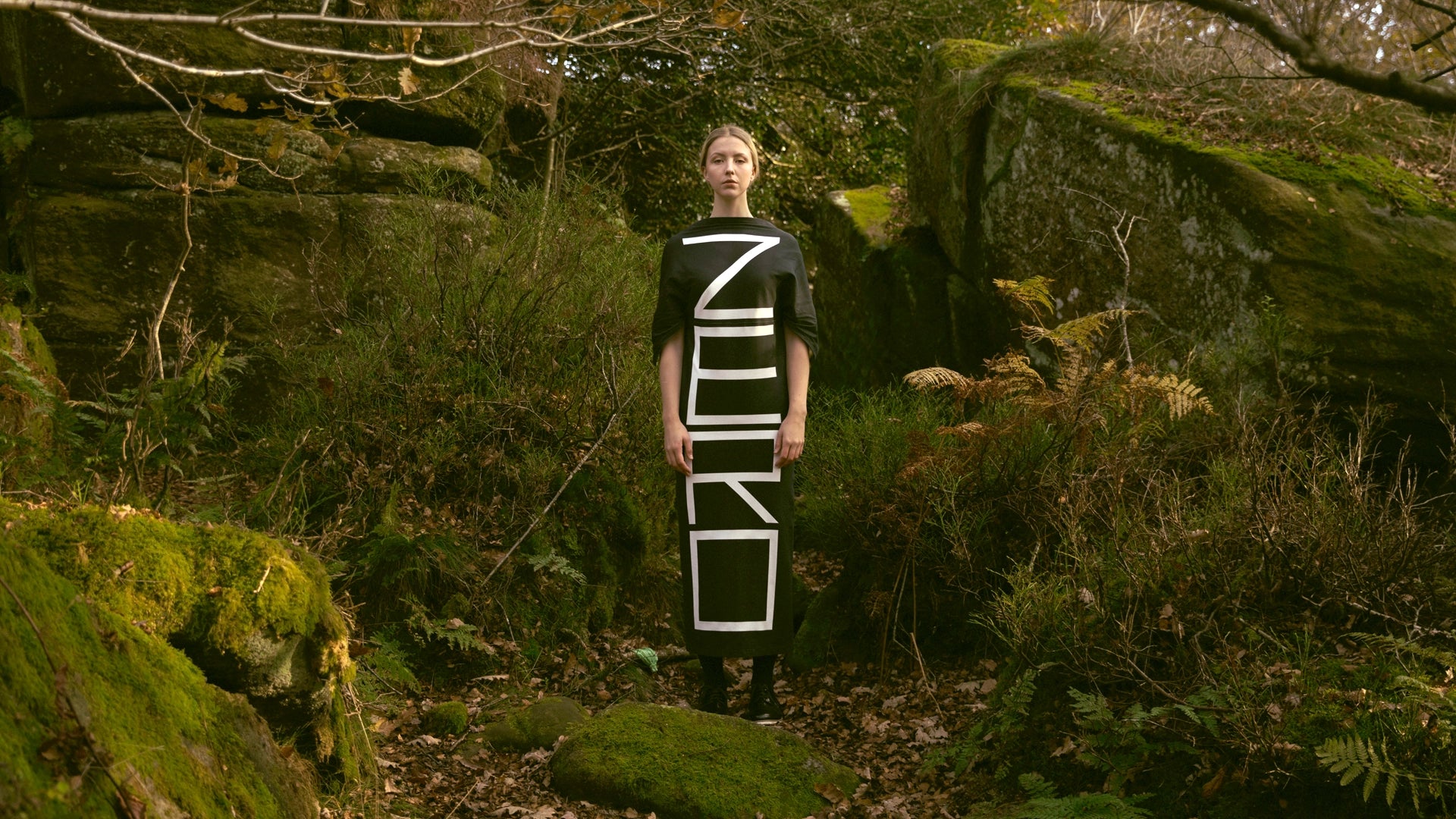 A model stands among moss-covered rocks in a woodland setting, wearing a black dress printed with the word ZERO, embodying Cunnington & Sanderson's zero-waste design philosophy.
